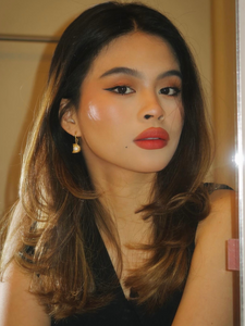 A young woman with long, wavy brown hair and brown eyes, wearing a black top and gold-colored clip-on hoop earrings with a floral design and central pearl accent. Her makeup includes winged eyeliner and bold red lipstick, and she has a serene and confident expression.