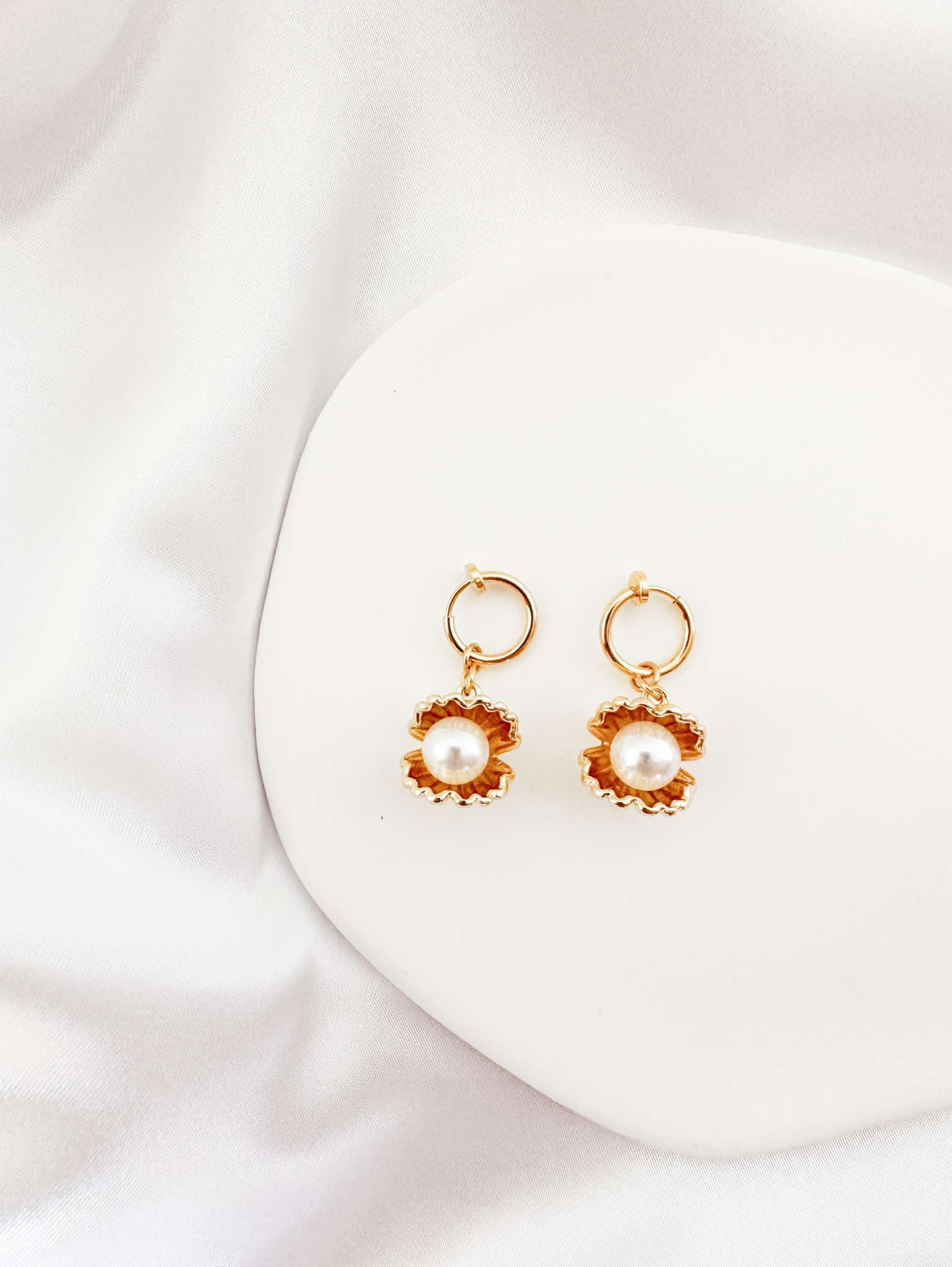 A pair of elegant gold-tone clip-on earrings with a scalloped, flower-like setting showcasing a central white pearl, presented on a white surface.