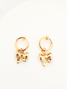 A pair of gold-colored clip-on earrings with delicate bow-shaped charms hanging from small circular hoops, set against a plain white background.