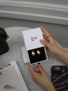 Gold earrings in a white box with 'jyc' branding, held by hands on a light surface.