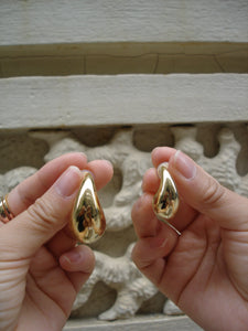 Two gold teardrop-shaped rings held by a hand against a textured wall.