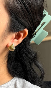 Close-up of an ear wearing a gold shell-shaped earring with dark hair and a light gray shirt.