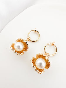 A pair of elegant gold-tone clip-on earrings with a scalloped, flower-like setting showcasing a central white pearl, presented on a white surface.