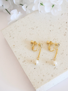 These screwback clip-on earrings feature a gold plated metal base with a cubic zirconia charm hanging from a short dangle pearl. The earrings are designed to be worn without the need for pierced ears. The screwback mechanism ensures a secure and comfortable fit for all-day wear.