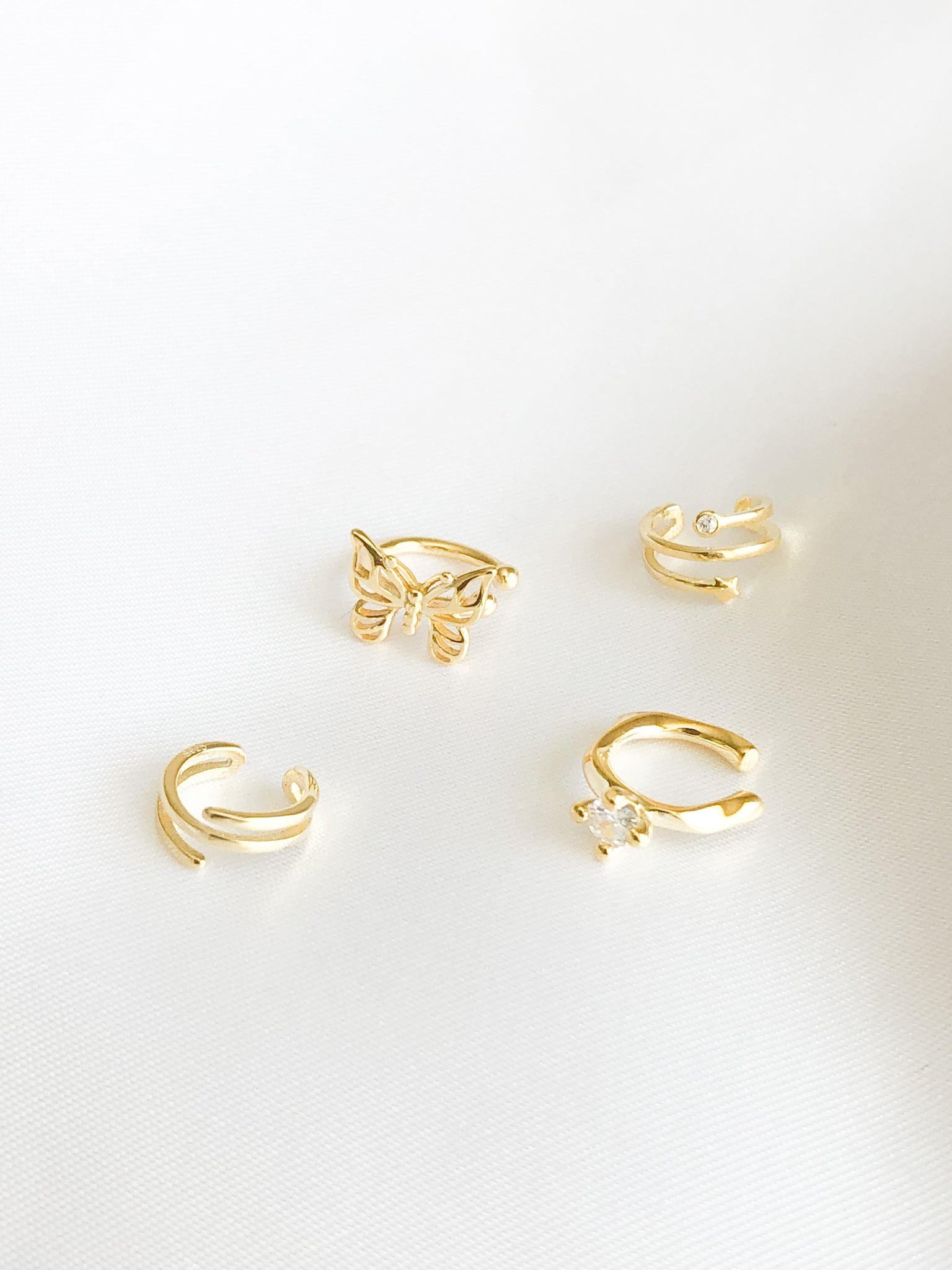 Upgrade your jewelry game with our stunning bundle of gold ear cuffs, featuring a gold butterfly ear cuff, a gold 3 banded layered ear cuff, a gold 3 banded layered ear cuff with star and one gem, and a gold cuff with cubic zirconia gem. All cuffs are designed for a comfortable and secure fit, perfect for those without pierced ears. 